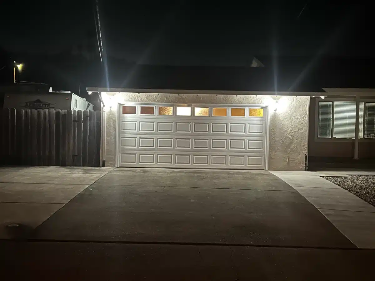 A white traditional raised-panel garage door with illuminated top windows glows under exterior lights at night, emphasizing the security provided by professional garage door repair.