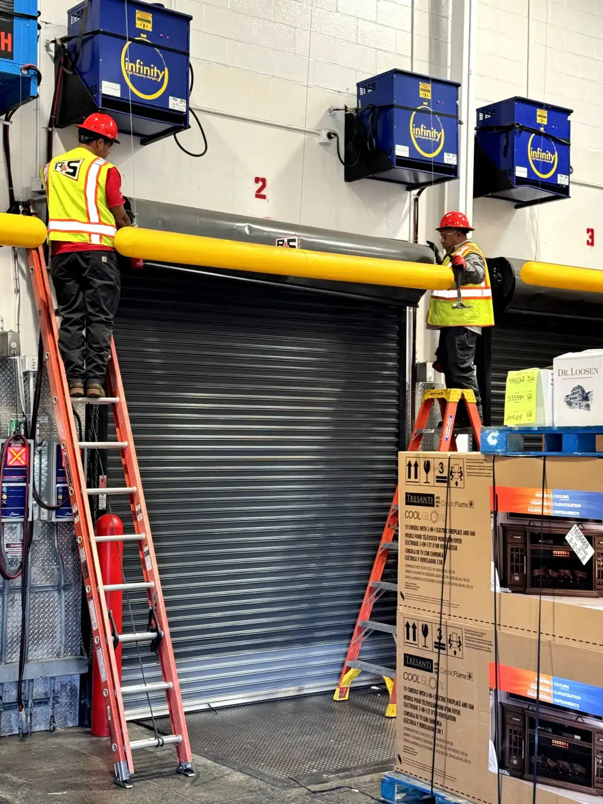 Two technicians wearing safety vests and hard hats work from ladders to service a heavy-duty industrial roll-up dock door, demonstrating the specialized care required for commercial overhead doors.