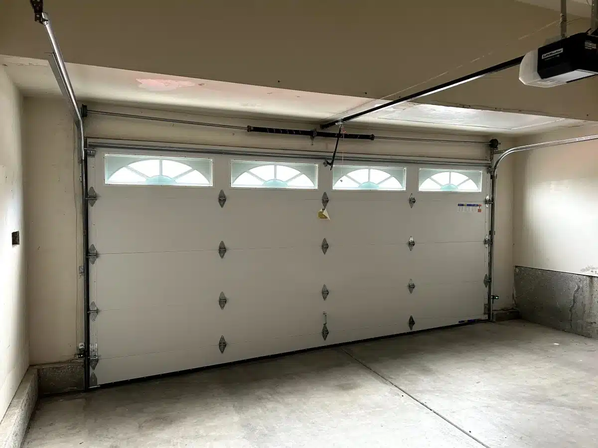 An interior view shows a white sectional door with arched sunray top windows and an overhead spring system, highlighting a completed garage door torsion spring replacement.