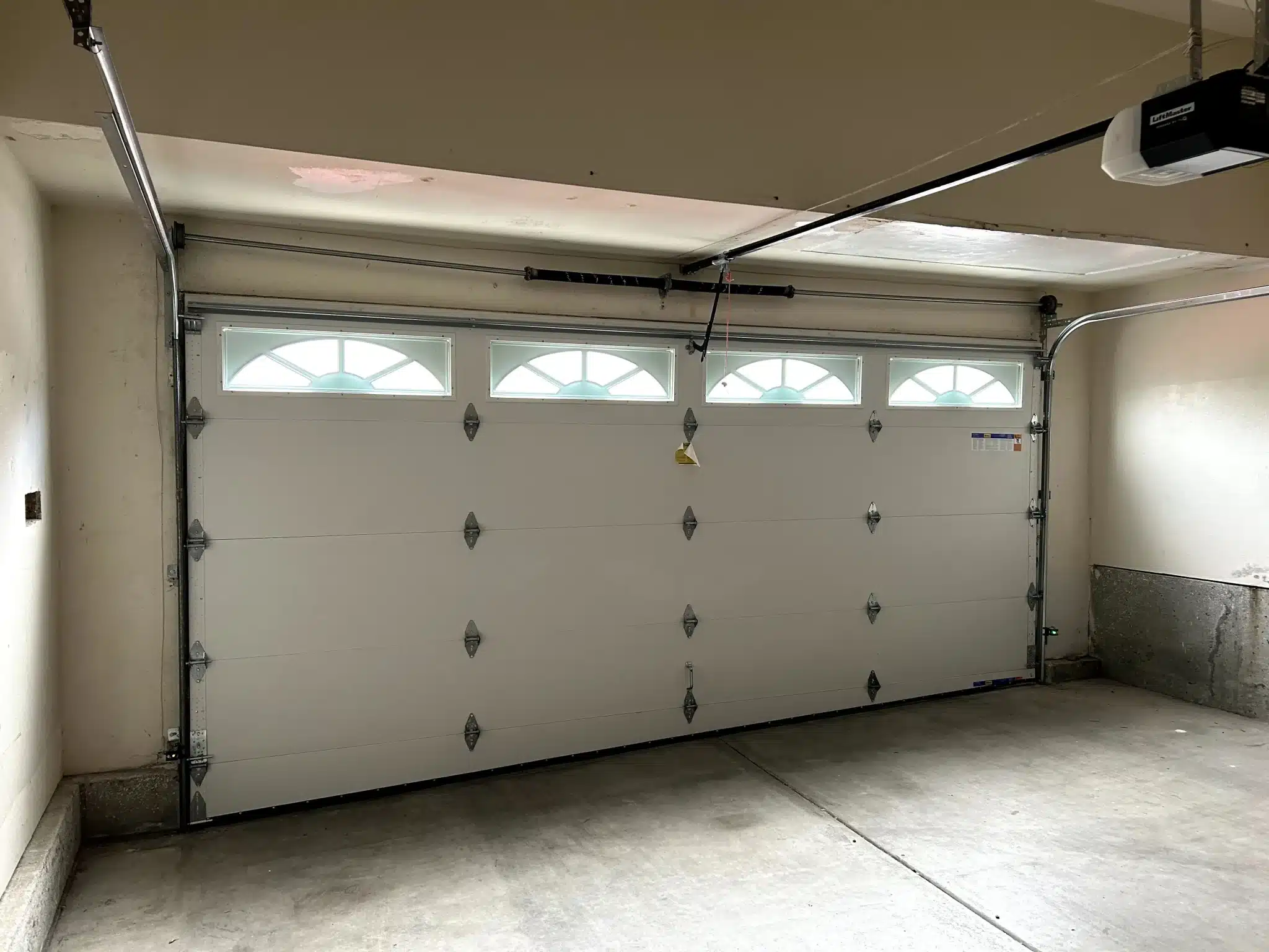 An interior view shows a white sectional door with arched sunray top windows and an overhead spring system, highlighting a completed garage door torsion spring replacement.