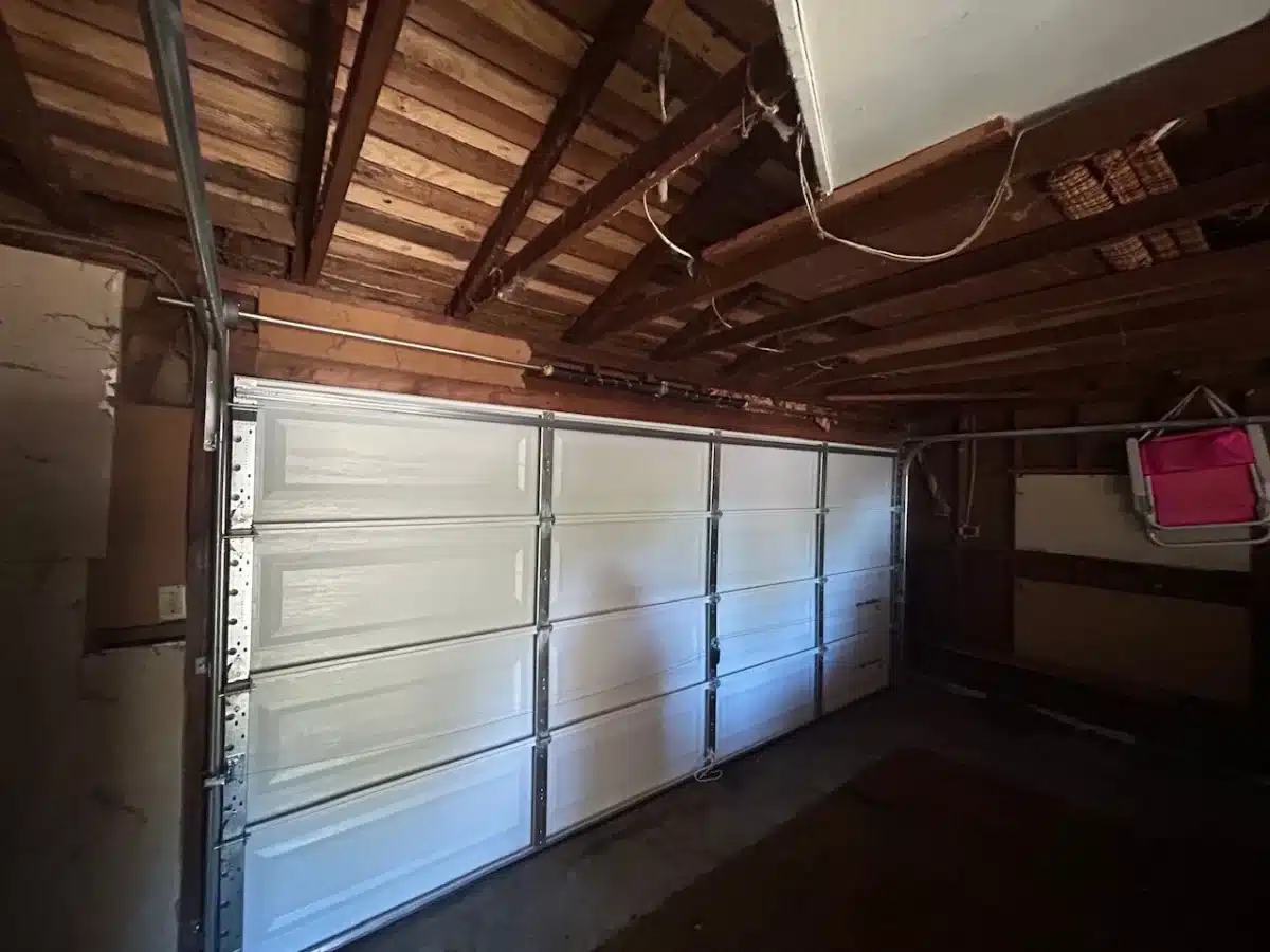 An interior perspective shows a white sectional garage door closed beneath a rustic wooden ceiling structure, highlighting the need to check components early for common garage door spring warning signs.