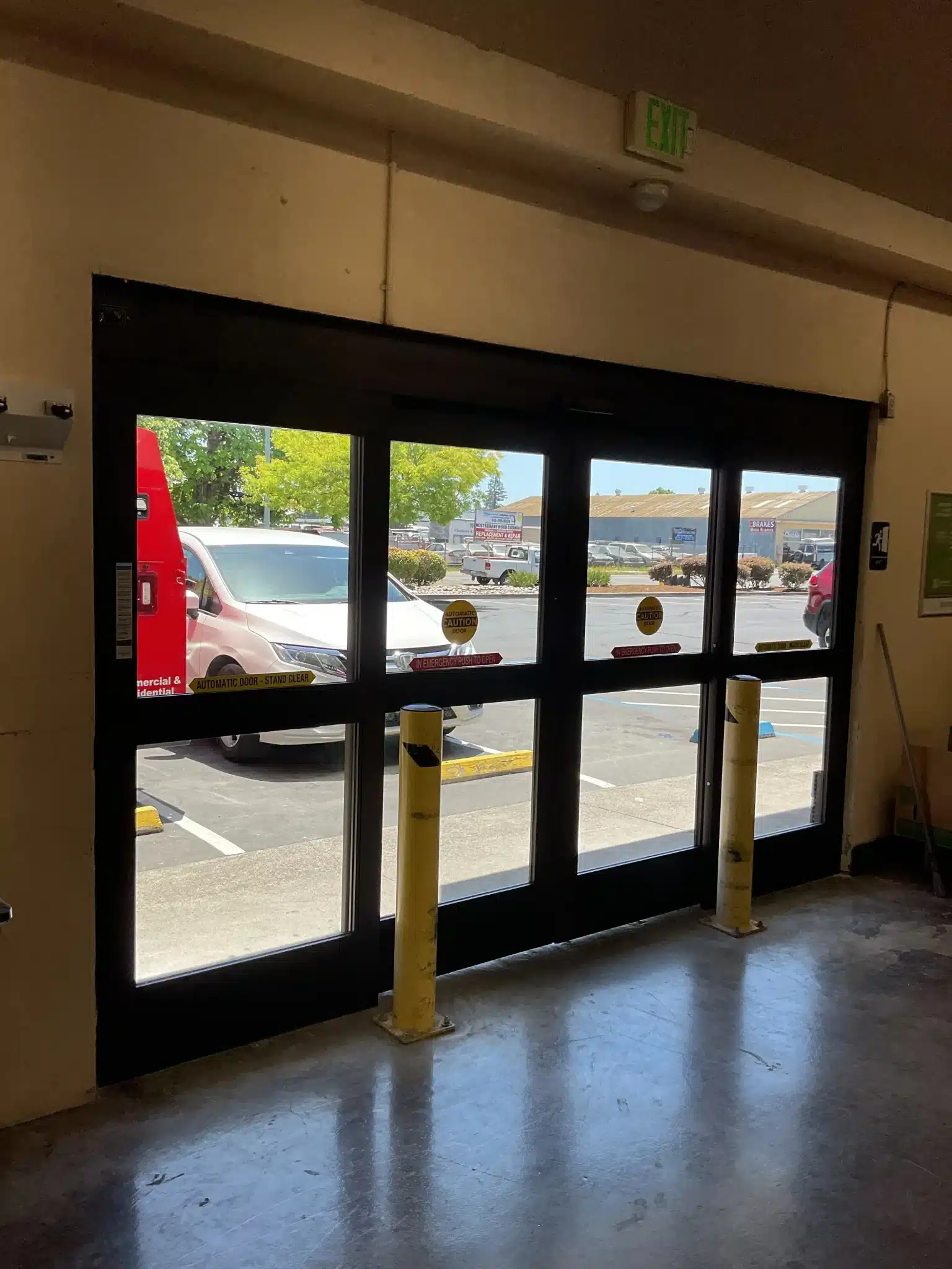 An interior view looks out through a dark-framed sliding glass entryway guarded by yellow safety bollards, demonstrating the accessibility and convenience provided by commercial automatic door benefits.